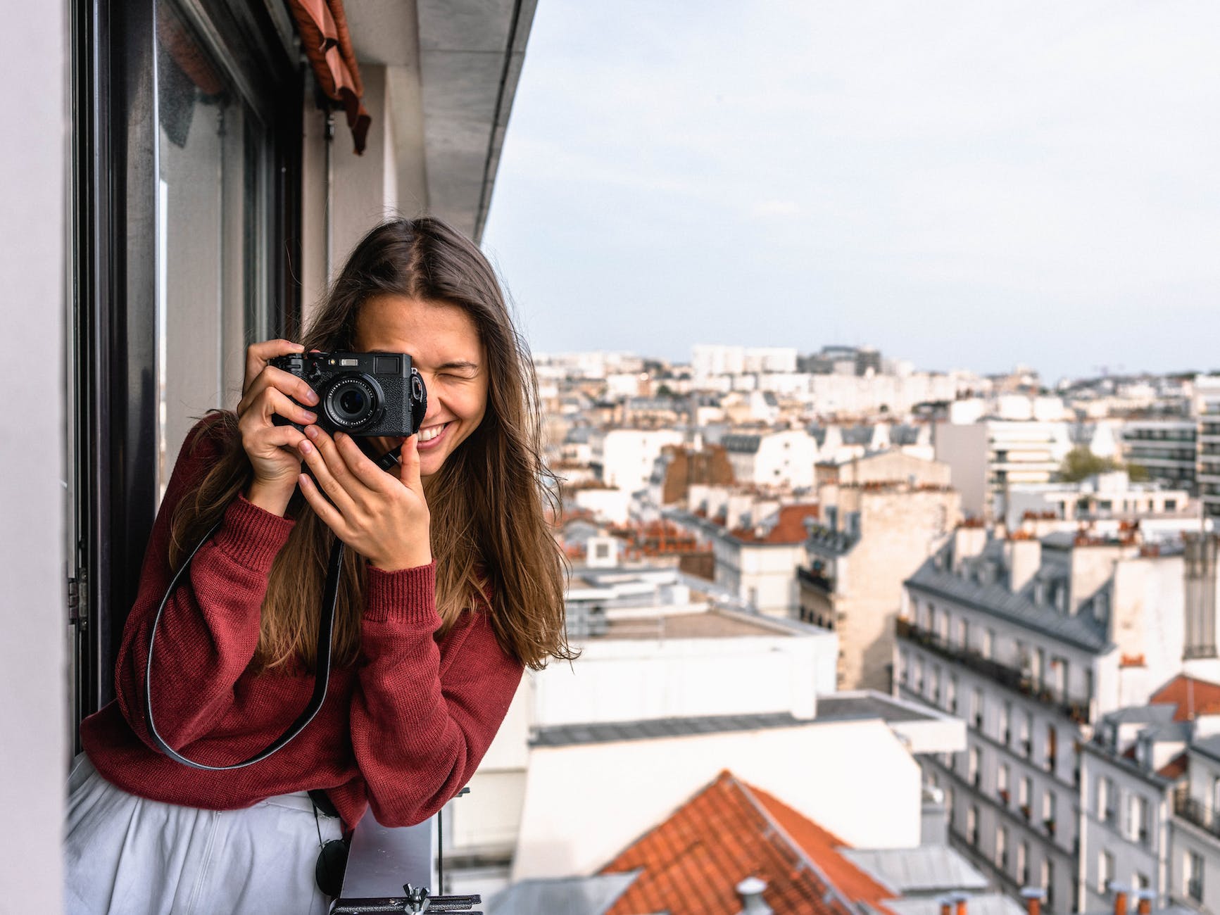 woman wearing maroon sweater standing on veranda using camera while smiling overlooking houses and buildings