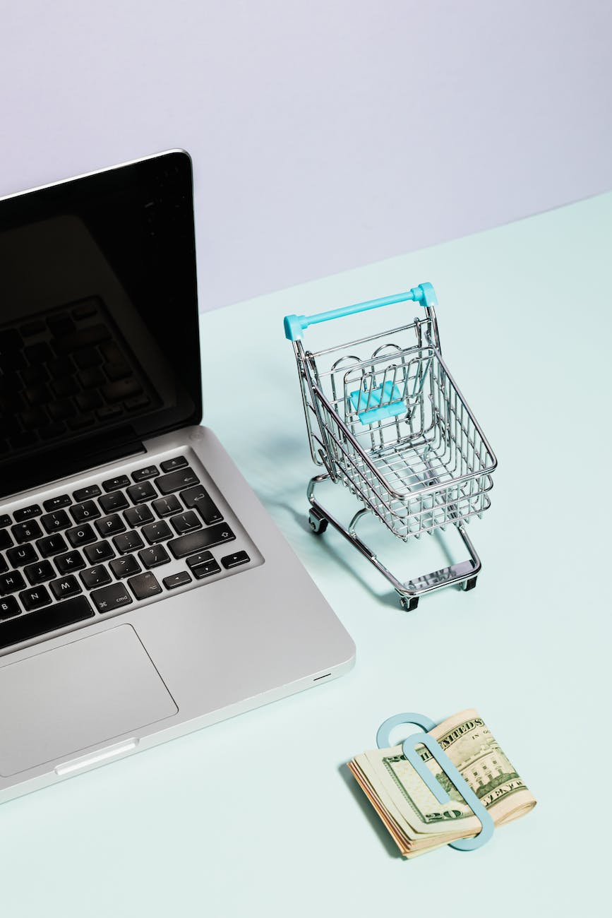 a macbook beside a miniature shopping cart and clipped money